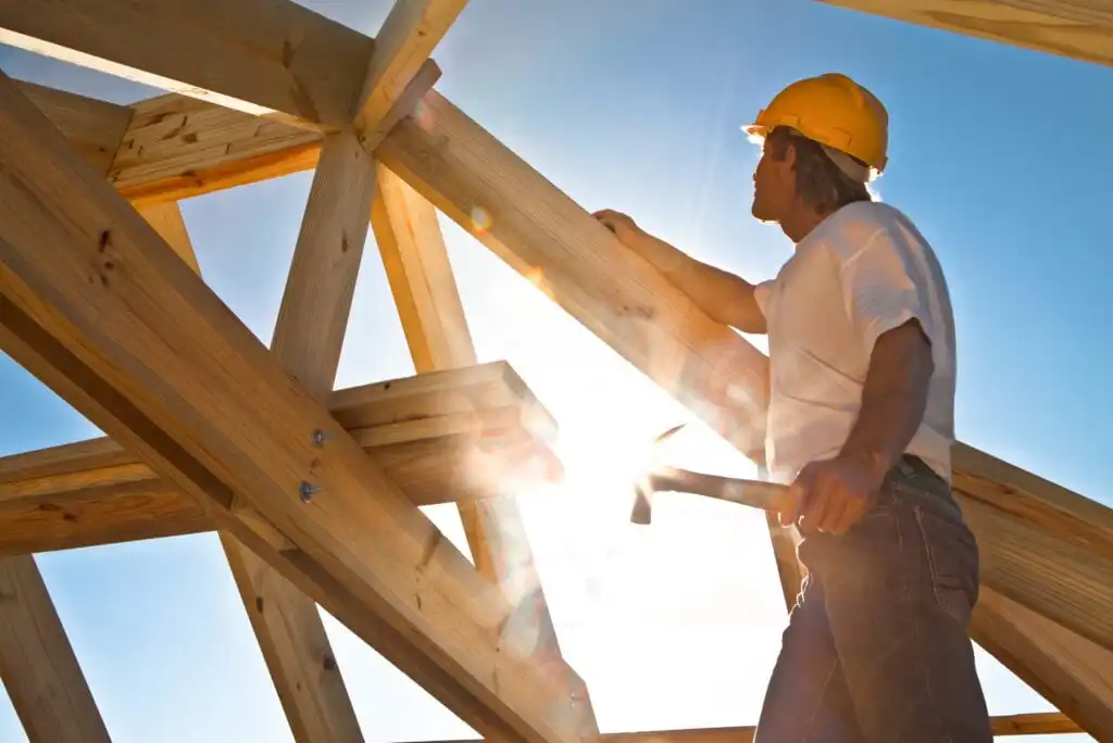 A construction worker wearing a yellow hard hat and white shirt holds a hammer while working on a wooden structure under bright sunlight and a clear blue sky.
