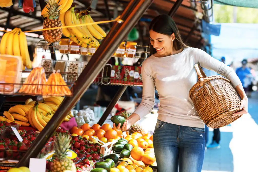 A woman holding a wicker basket smiles while selecting fresh produce at an outdoor market stand filled with fruits like bananas, oranges, strawberries, and avocados on a sunny day.