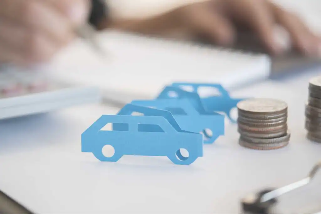 Three blue paper car cutouts are on a desk near stacks of coins, with a hand using a calculator and writing in a notebook in the background, suggesting car finances or budgeting.