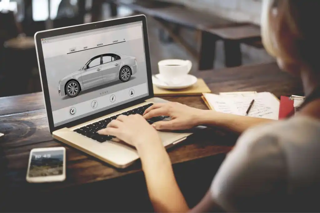 A person using a laptop at a wooden table, viewing a car customization or shopping website. A cup of coffee, notepad with pen, and smartphone are also on the table.