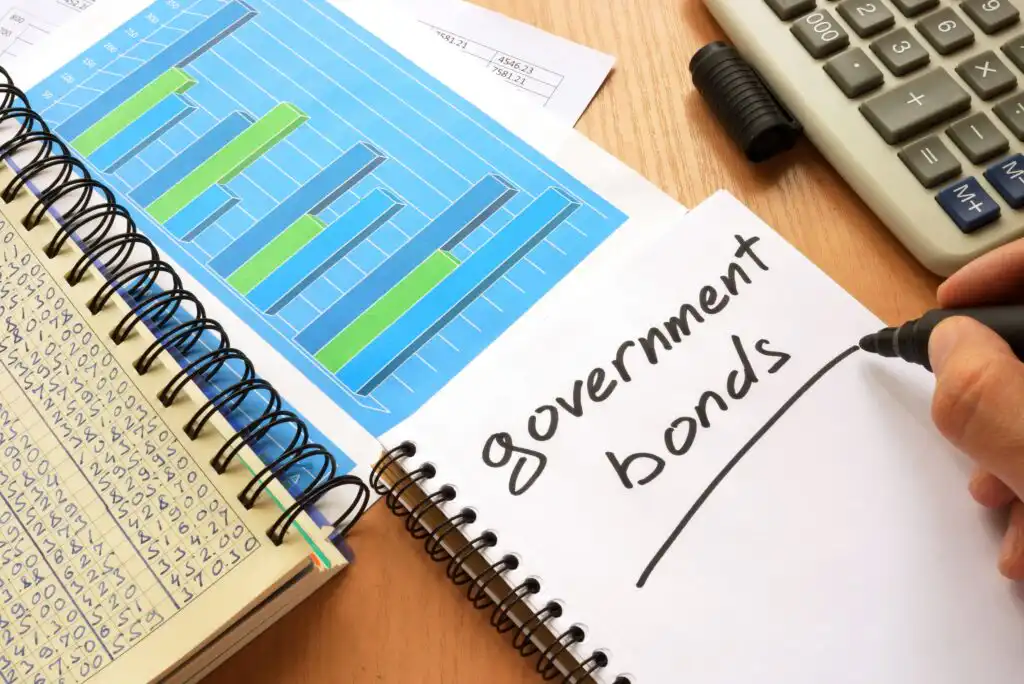 A person writes “government bonds” in a notebook beside a chart with bar graphs, another notebook with numbers, a calculator, and a pen on a wooden desk.