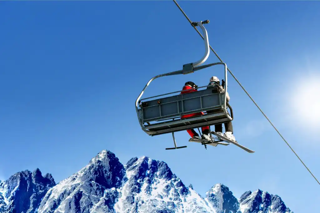 Two skiers ride a ski lift above snowy mountain peaks under a clear blue sky, with the sun shining brightly in the background.