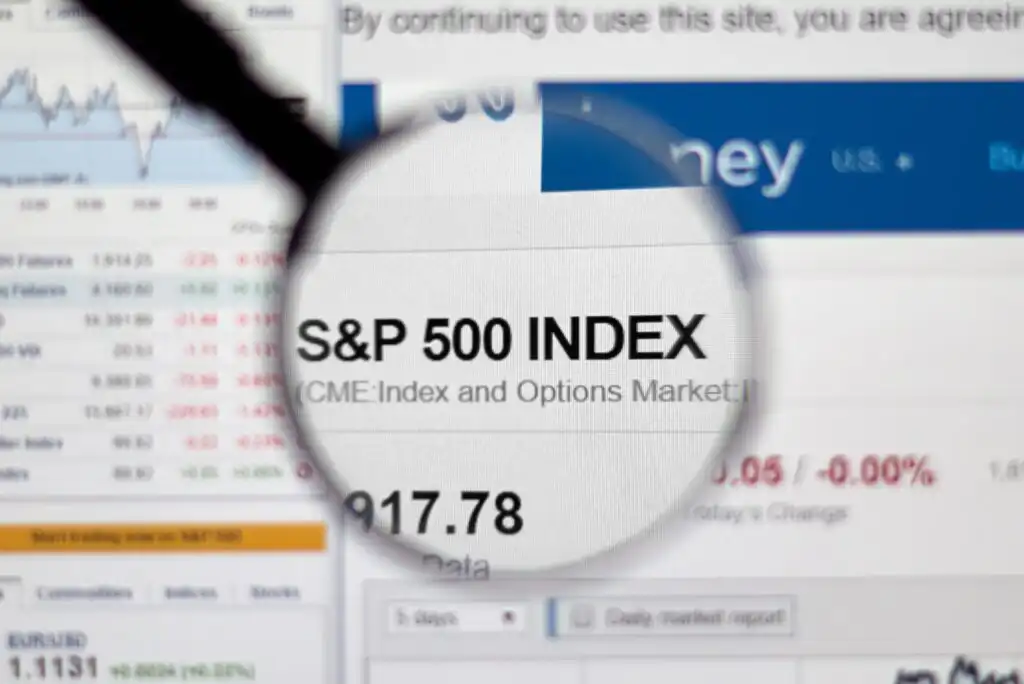 A magnifying glass highlights the text S&P 500 INDEX on a computer screen displaying financial data and stock market charts in the background.