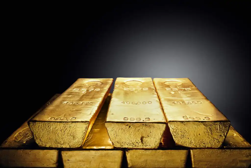 Three large gold bars are stacked side by side on top of more gold bars against a dark gradient background, showcasing their shiny, reflective surfaces and engraved markings.