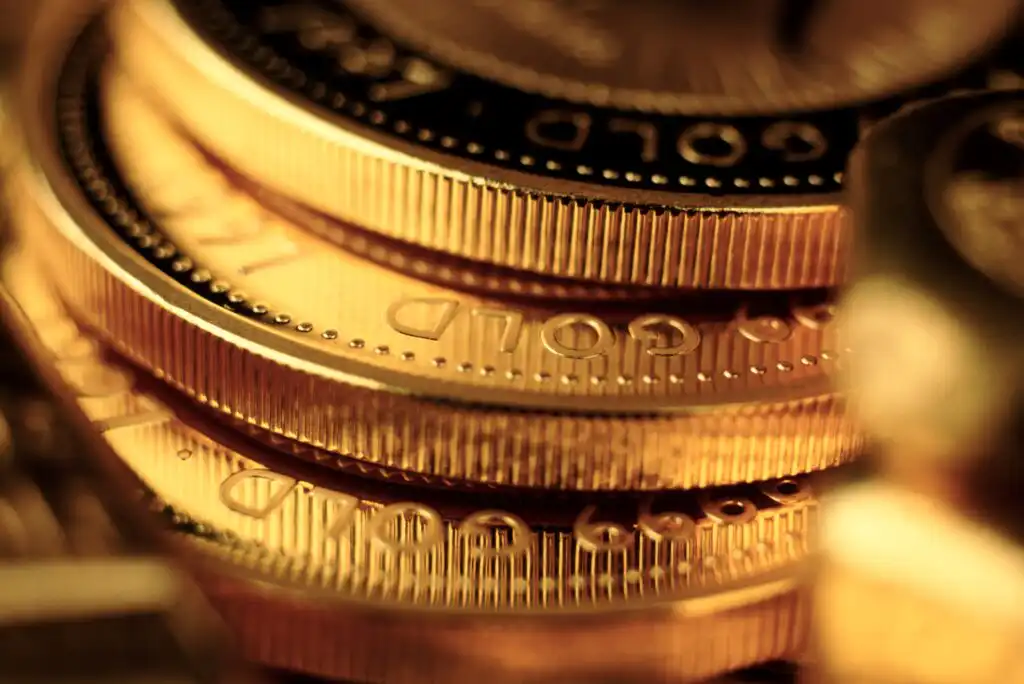 A close-up of stacked gold coins, showing detailed ridges and the word GOLD engraved on the edges, with a warm, golden glow highlighting the metallic surfaces.