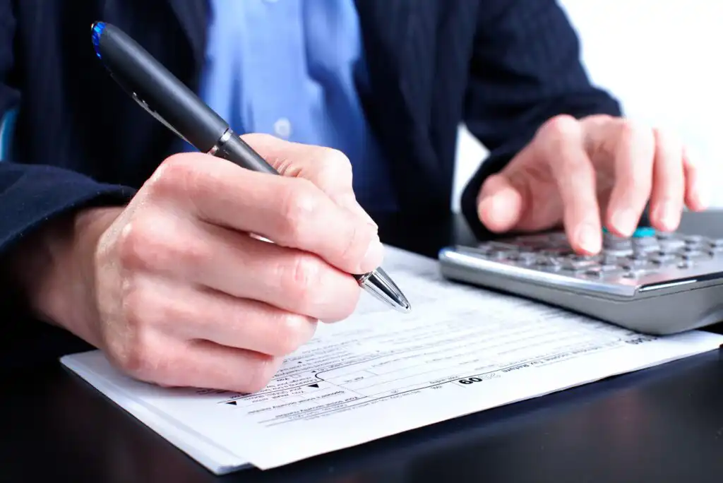 A person in business attire fills out a form with a pen while using a calculator, suggesting they are working on financial or tax documents at a desk.