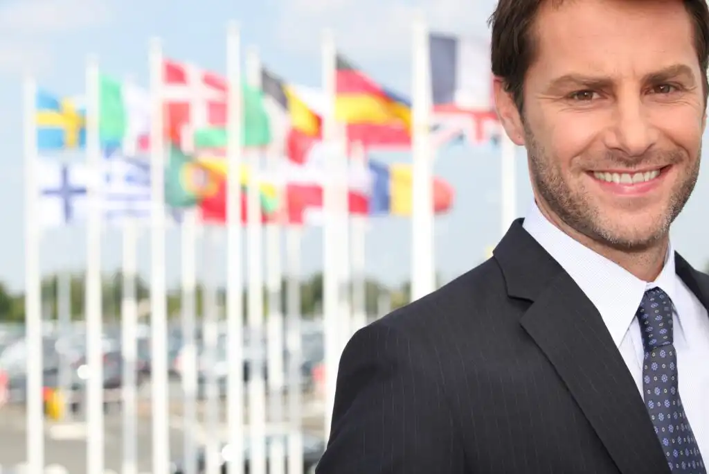 Smiling man in a suit stands outdoors with several blurred international flags in the background on a sunny day.