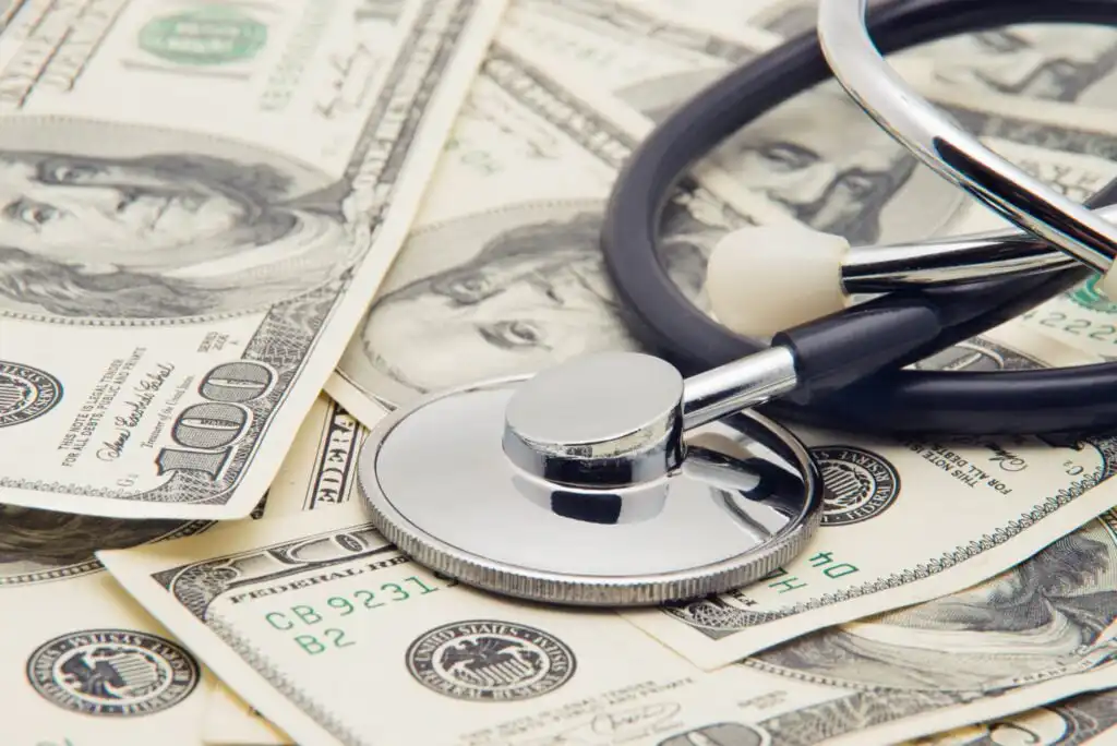 A stethoscope rests on top of several scattered U.S. hundred-dollar bills, symbolizing the connection between healthcare and financial costs.