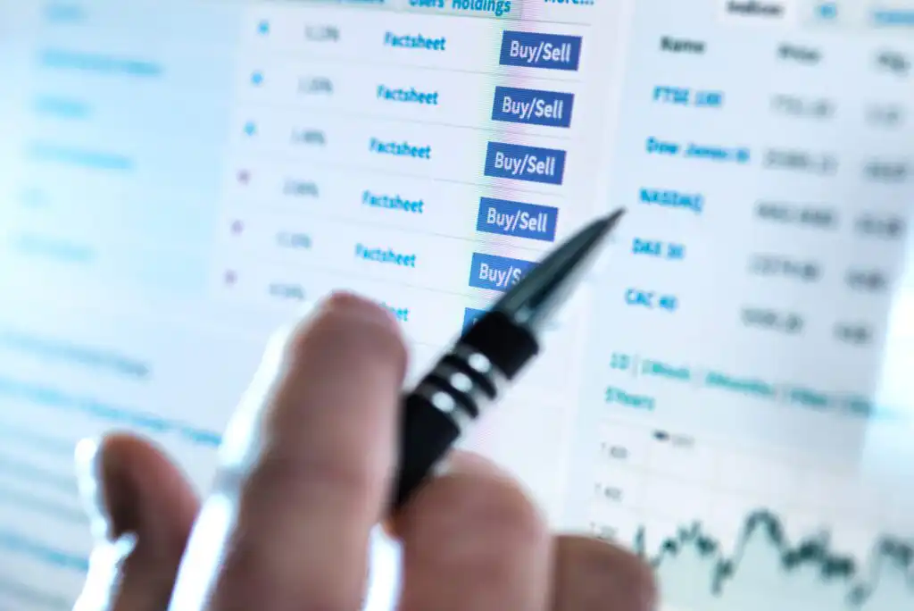 A close-up of a hand holding a pen and pointing at a computer screen displaying financial data, including stock market factsheets and blue Buy/Sell buttons. A graph is visible in the lower right corner.