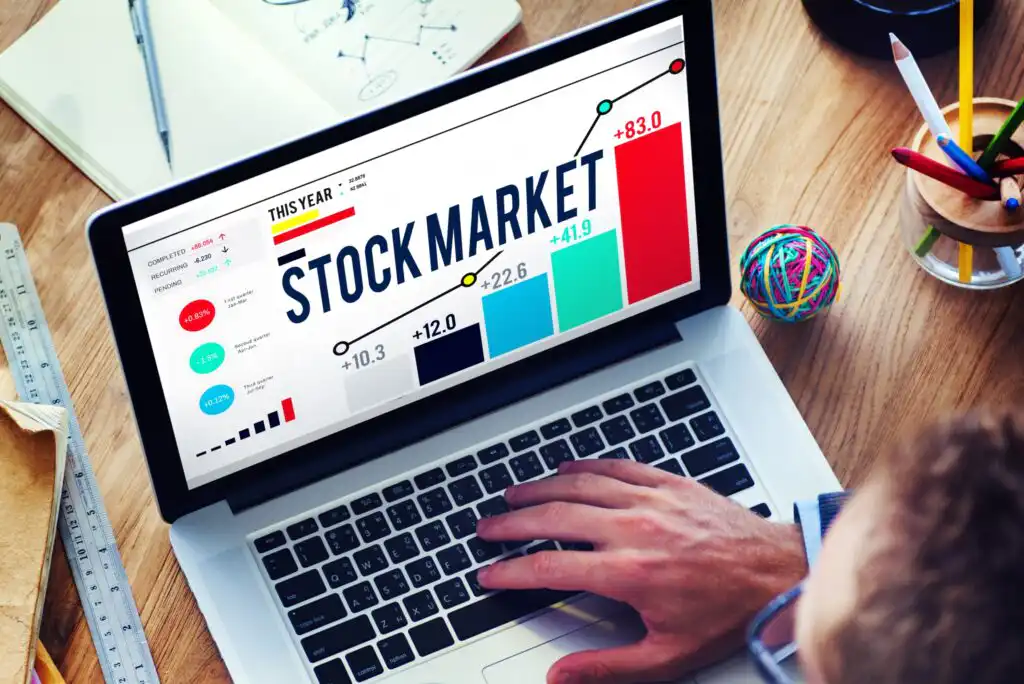 A person uses a laptop displaying a colorful stock market chart with bars and numbers, showing market trends and a STOCK MARKET heading. Other office items are visible on the wooden desk.