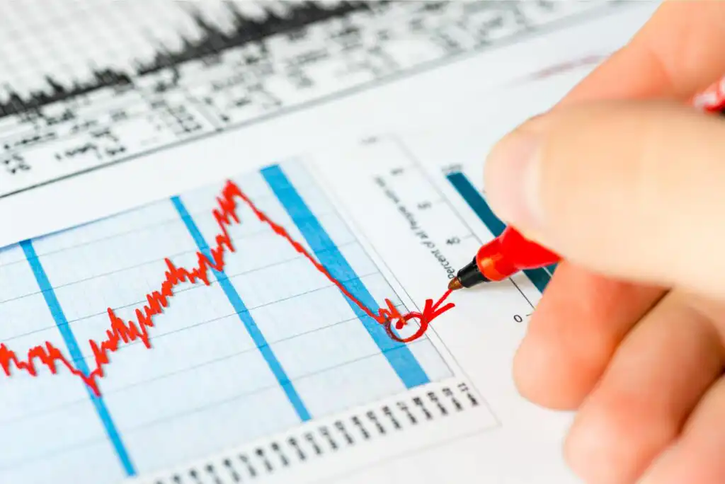 A hand holding a red pen circles a significant dip on a printed financial chart with a red line graph, indicating a drop and partial recovery in data, with more charts visible in the background.