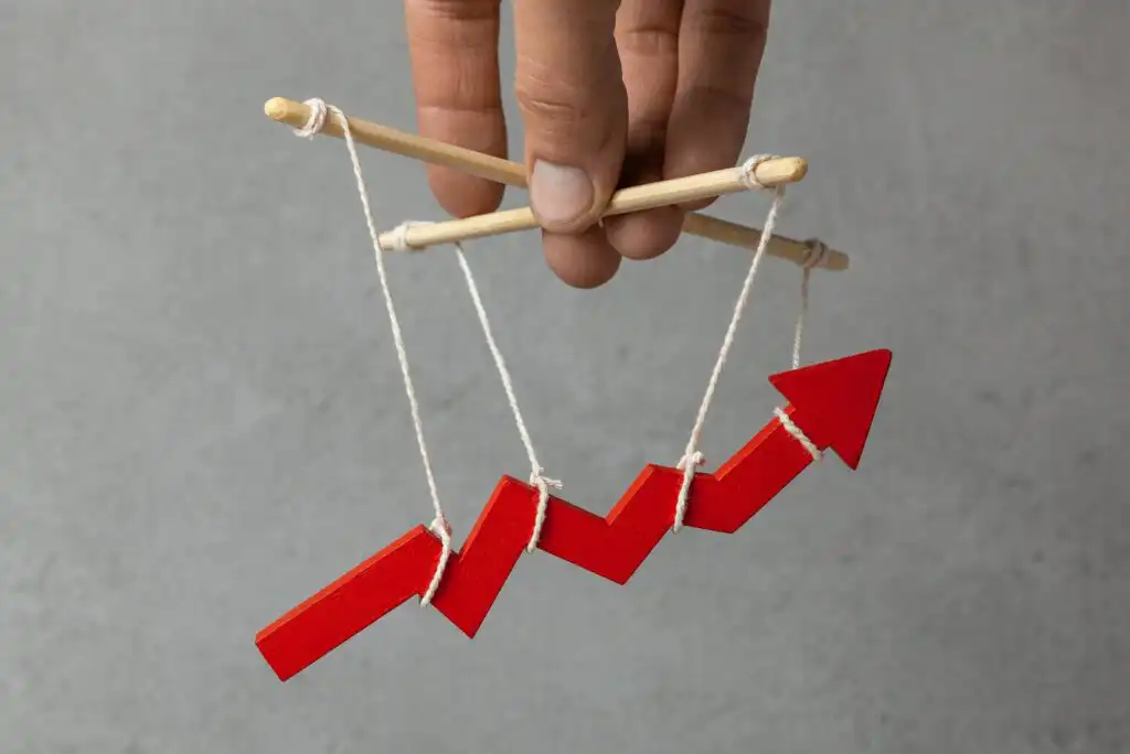 A hand holds wooden sticks and strings like a puppet, controlling a red, upward-pointing zigzag arrow, symbolizing manipulation of financial or business growth. Gray background.