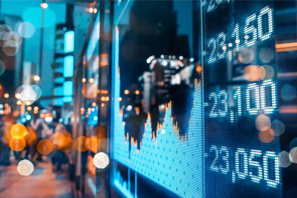 A digital stock market display shows fluctuating numbers and graphs at night, with city lights and blurred figures of people walking on a busy urban street in the background.