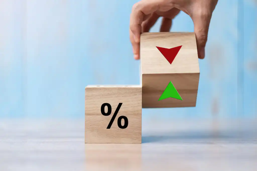 A hand adjusts wooden blocks: one block shows a percent sign, while the other displays a red down arrow and a green up arrow, symbolizing fluctuating interest or percentage rates.