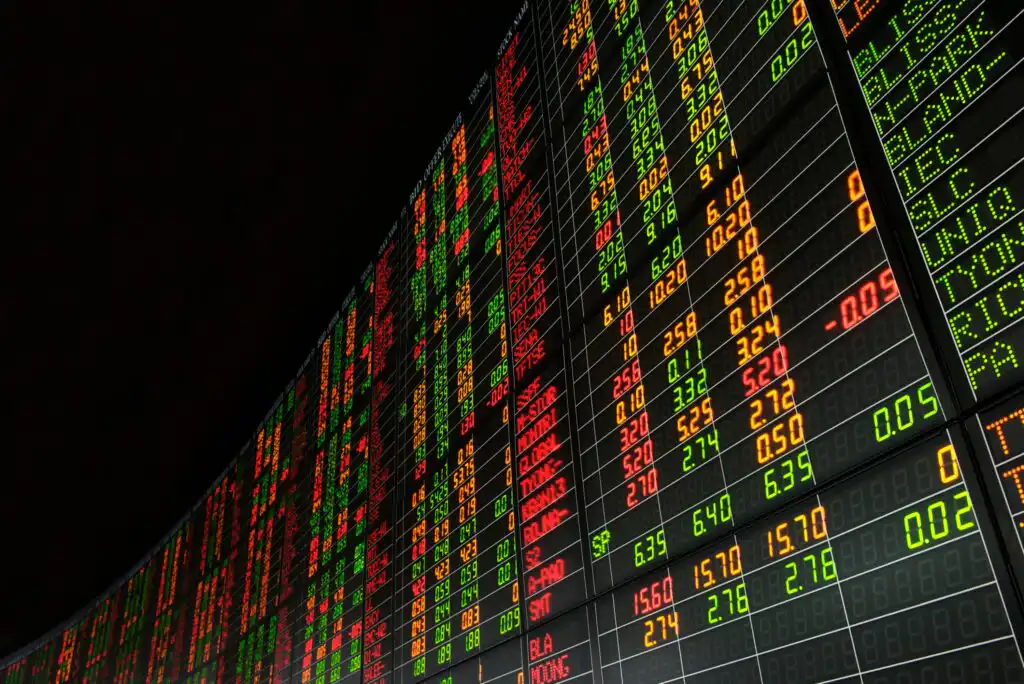 A large digital stock market board displays rows of numbers and letters in red, green, yellow, and white, indicating fluctuating financial data on a black background.