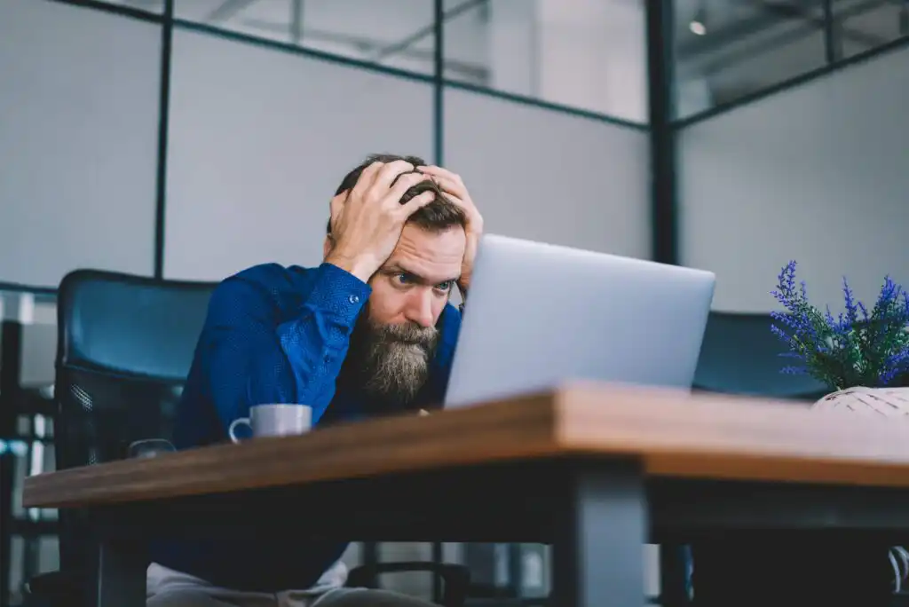 A man with a beard wearing a blue shirt sits at a desk, holding his head in frustration while staring at a laptop screen in a modern office setting. A cup and a potted plant are on the desk.