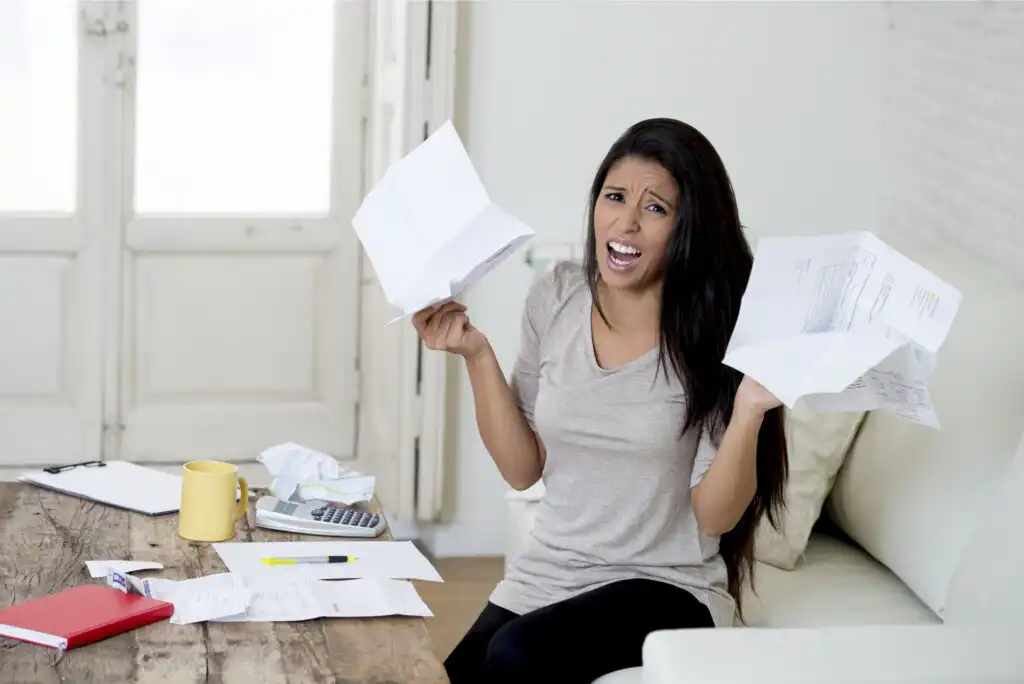 A woman sits on a couch holding crumpled papers, looking frustrated. A calculator, coffee mug, and scattered documents are on the wooden table in front of her. She appears stressed about bills or finances.