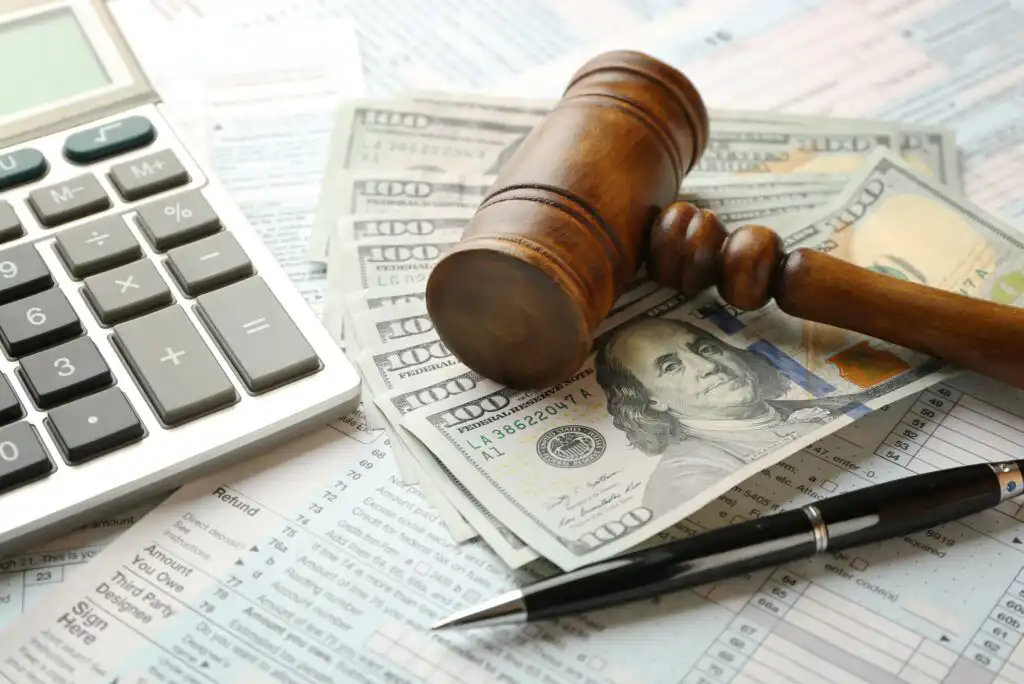 A wooden gavel rests on a stack of hundred-dollar bills, with a pen and a calculator nearby, all placed on tax forms, symbolizing financial or legal decisions.
