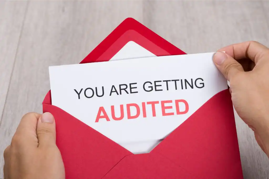 A person pulls a letter from a red envelope. The letter reads YOU ARE GETTING AUDITED with AUDITED highlighted in red text. The background is a light wood surface.