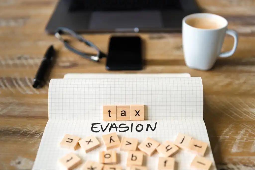 A notepad with the words tax evasion spelled out using letter tiles sits on a wooden desk, surrounded by a pen, smartphone, eyeglasses, a cup of coffee, and a laptop in the background.
