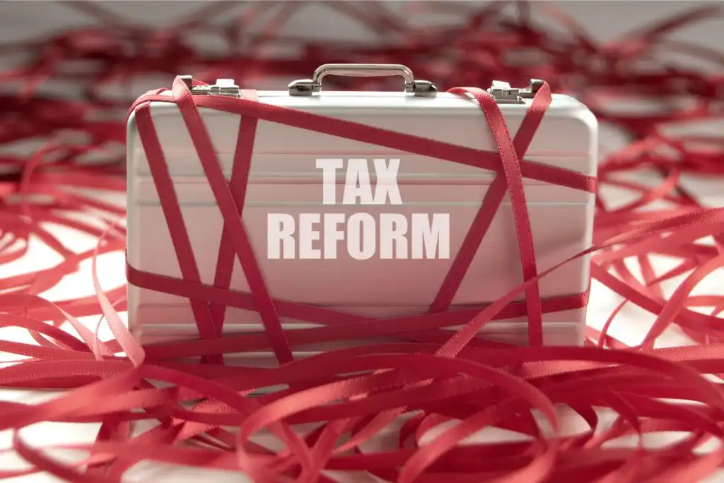 A silver briefcase labeled TAX REFORM is wrapped in red tape, with more red tape scattered around, symbolizing bureaucratic hurdles and complications related to tax reform.