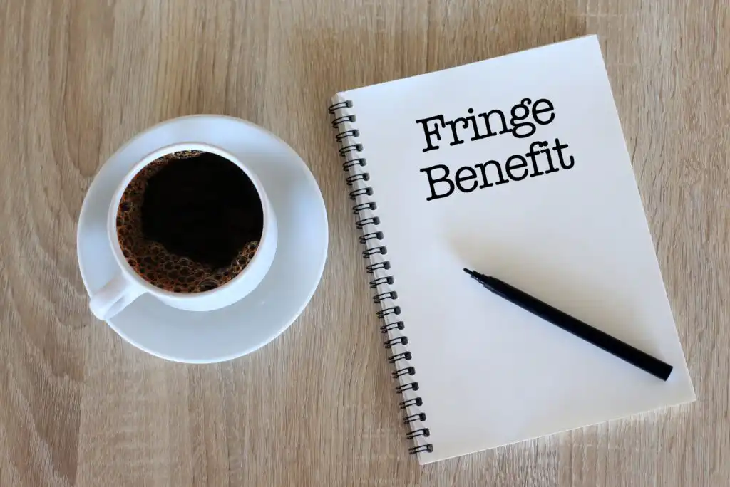 A cup of black coffee on a white saucer sits next to a spiral notebook with Fringe Benefit written on the page, and a black pen resting on the notebook, all on a wooden surface.