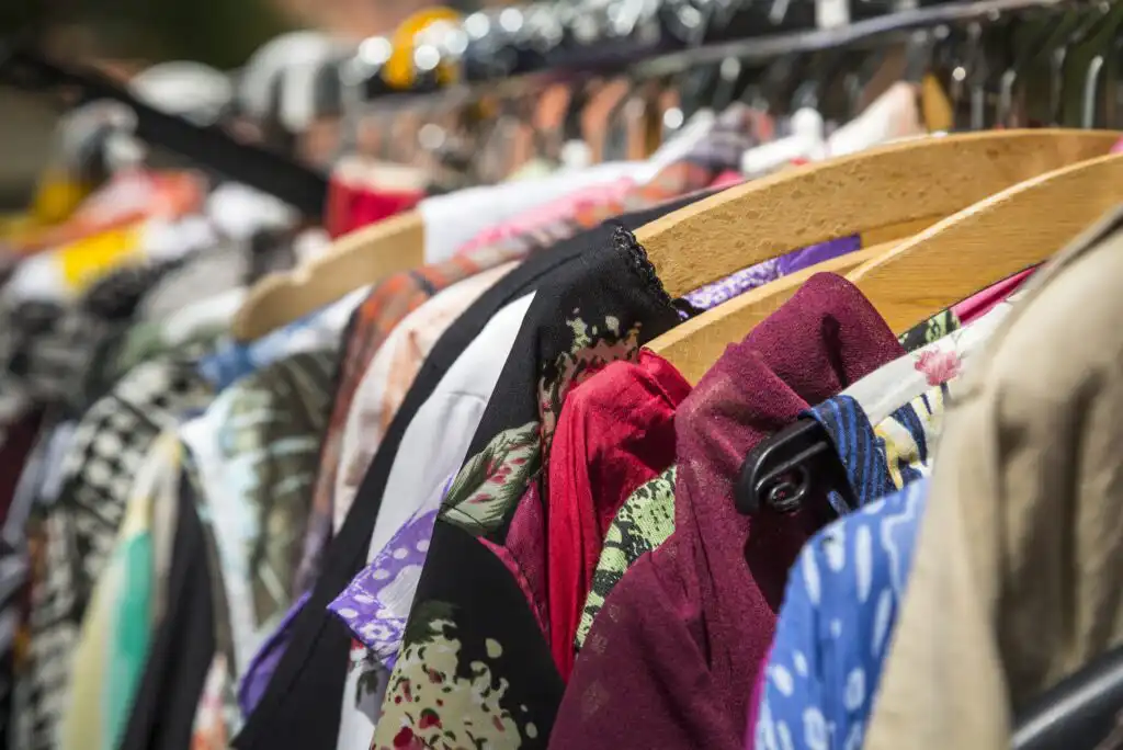 A close-up view of colorful clothing hanging on wooden and plastic hangers on a crowded clothes rack, displaying various patterns and fabrics in bright sunlight.