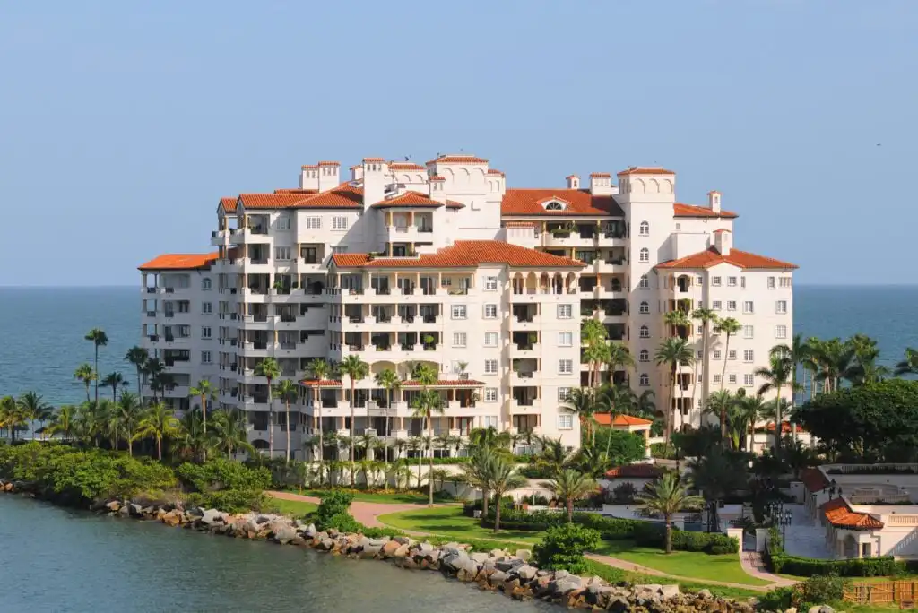 Large Mediterranean-style condominium building with red-tiled roofs, surrounded by palm trees, sits near the water’s edge with ocean in the background under a clear blue sky.