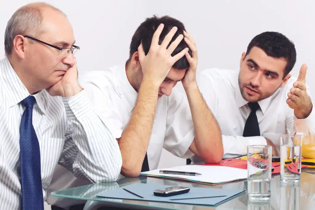 Three men in business attire sit at a glass table. One man holds his head in frustration, while another gestures as if explaining something. The third man looks on thoughtfully. Papers, a pen, and drinks are on the table.