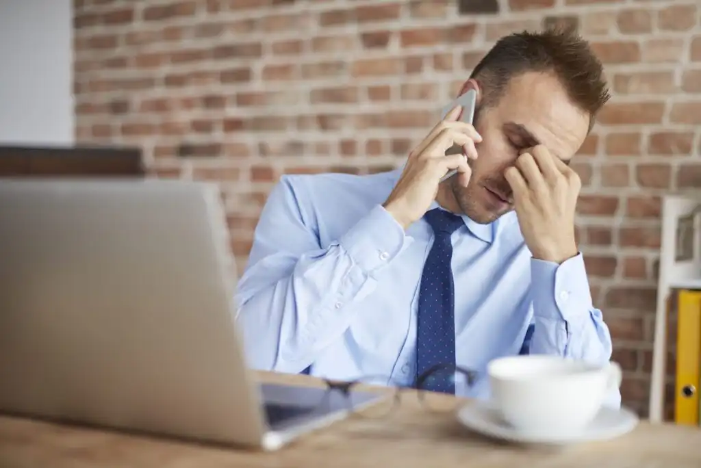 A man in a blue shirt and tie sits at a desk with a laptop, holding a phone to his ear and pinching the bridge of his nose, appearing stressed. A cup and saucer are on the table in front of him.