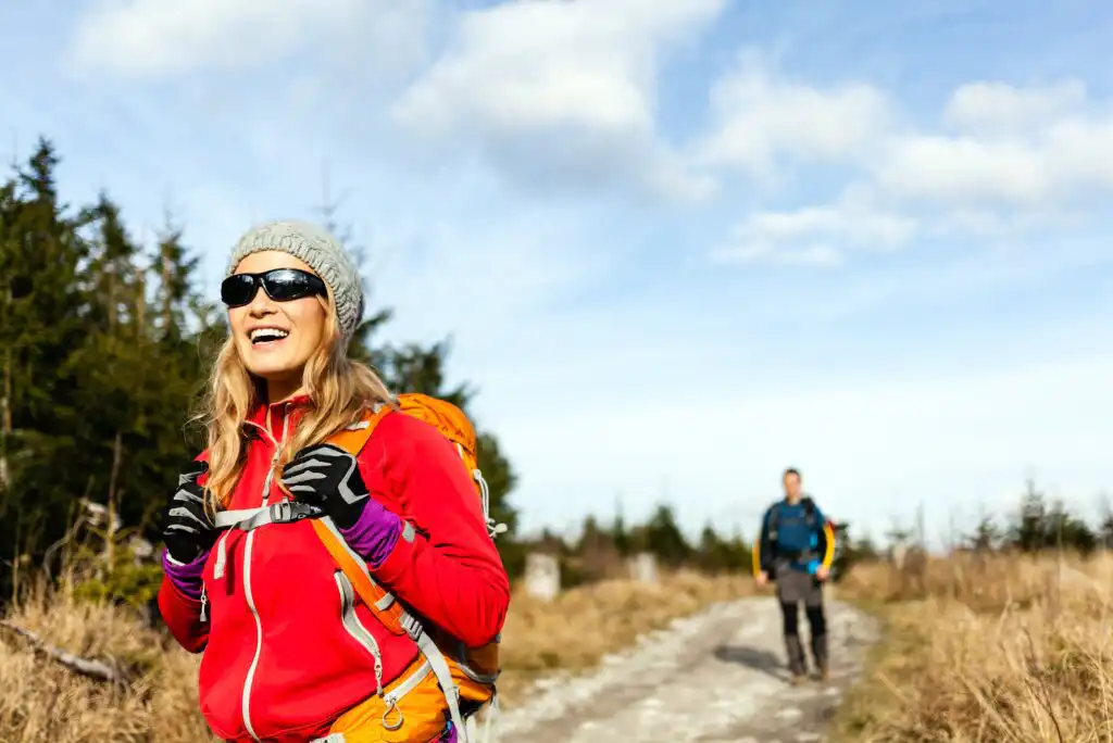 A woman wearing a red jacket, gray beanie, sunglasses, and gloves smiles while hiking on a trail with a backpack. A man walks behind her. The sky is partly cloudy, and trees line the path.