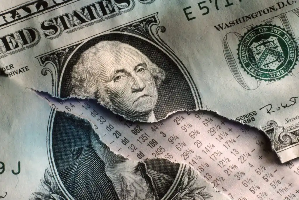 A close-up of a U.S. one-dollar bill with a jagged tear across George Washington’s portrait, revealing financial data printed on paper underneath.