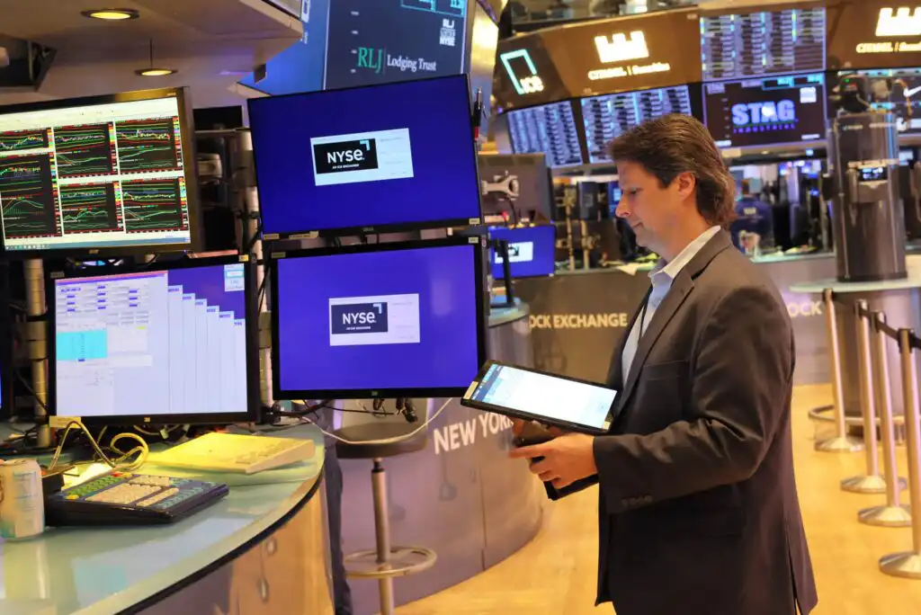 A man in a suit stands at a workstation with multiple monitors displaying NYSE information on a trading floor, holding a tablet and looking at the screens.