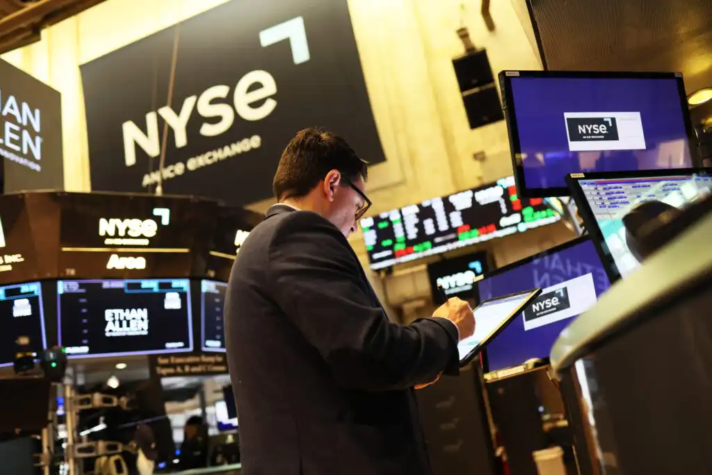 A person in a suit uses a tablet on the trading floor of the New York Stock Exchange, with electronic screens and the NYSE logo visible in the background.