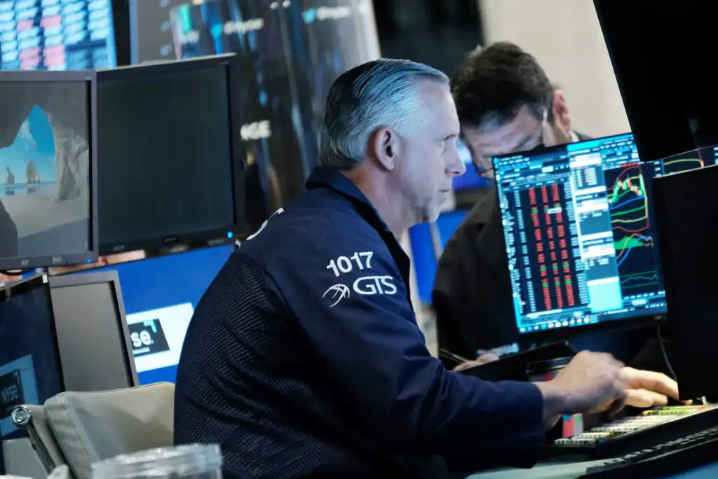 A man in a trading jacket works at a desk with multiple monitors displaying stock charts and market data. Another person is partially visible in the background, also focused on screens.