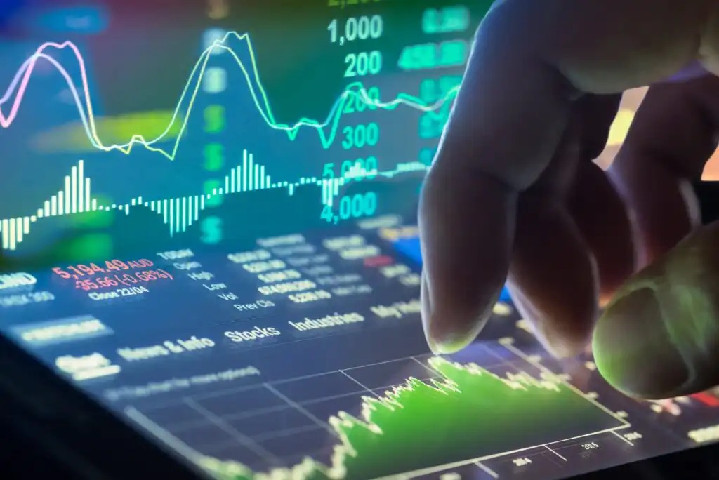 A close-up of a hand interacting with a digital tablet displaying colorful financial graphs, stock market charts, and data points in a technology-driven trading interface.