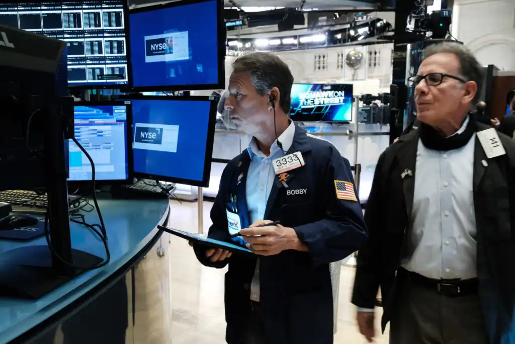 Two men stand on a trading floor; one holds a tablet and looks at computer monitors displaying stock data, while the other walks behind him. Multiple screens and a NYSE sign are visible in the background.