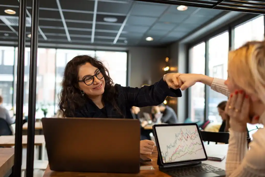 Two women sit at a table with laptops in a modern office. One woman, smiling, gives a fist bump to the other. A laptop screen shows a line graph with multiple colored lines.