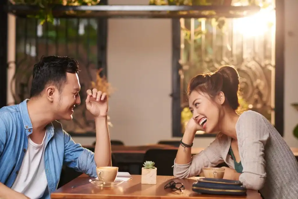 A man and woman sit at a café table, smiling and laughing together over coffee. Sunlight streams through the window behind them, creating a warm, cheerful atmosphere. A small plant and eyeglasses are on the table.
