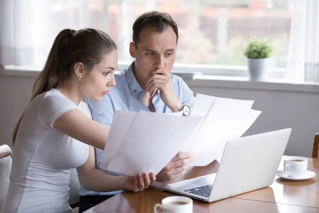 A worried couple sits at a table with paperwork and a laptop, reviewing documents together. The woman points at a paper while the man looks concerned, holding a sheet and resting his hand on his mouth.