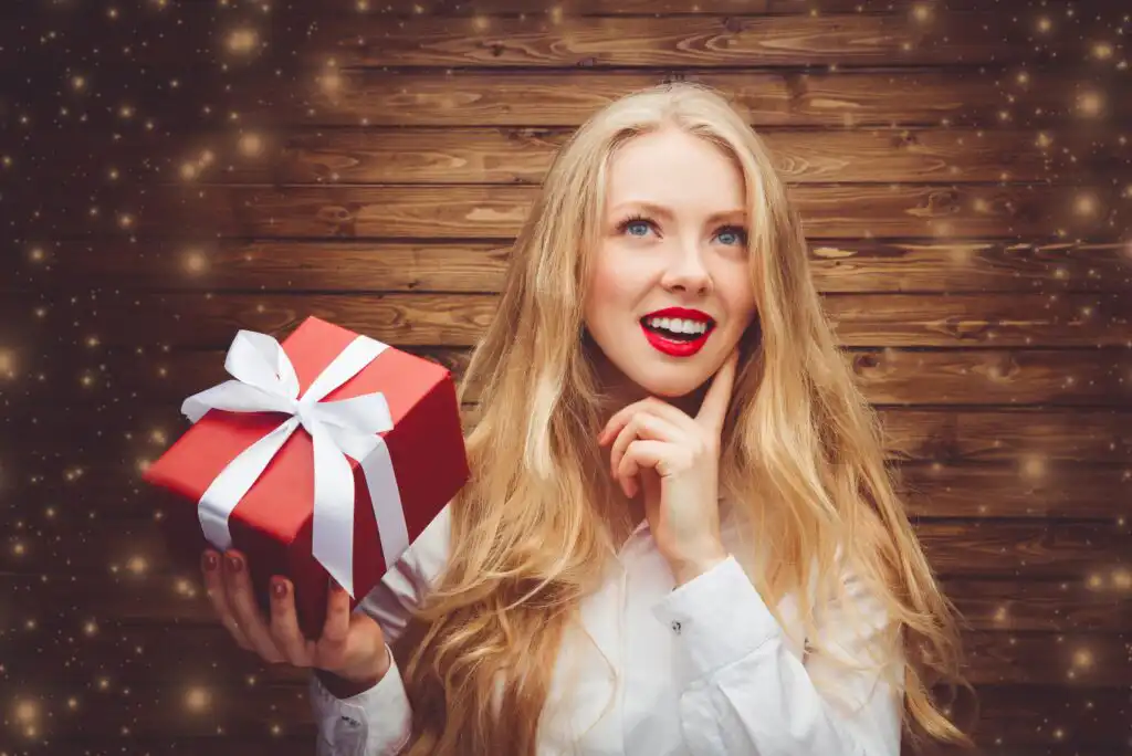 A smiling woman with long blonde hair holds a red gift box with a white ribbon against a wooden background, surrounded by sparkling lights. She looks thoughtful and happy, wearing a white shirt and red lipstick.