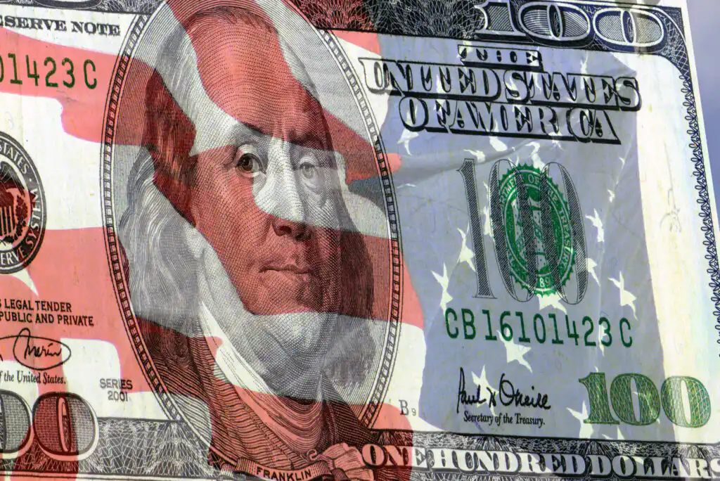 A U.S. 100 dollar bill featuring Benjamin Franklin is overlaid with a translucent American flag, blending currency and patriotic imagery.