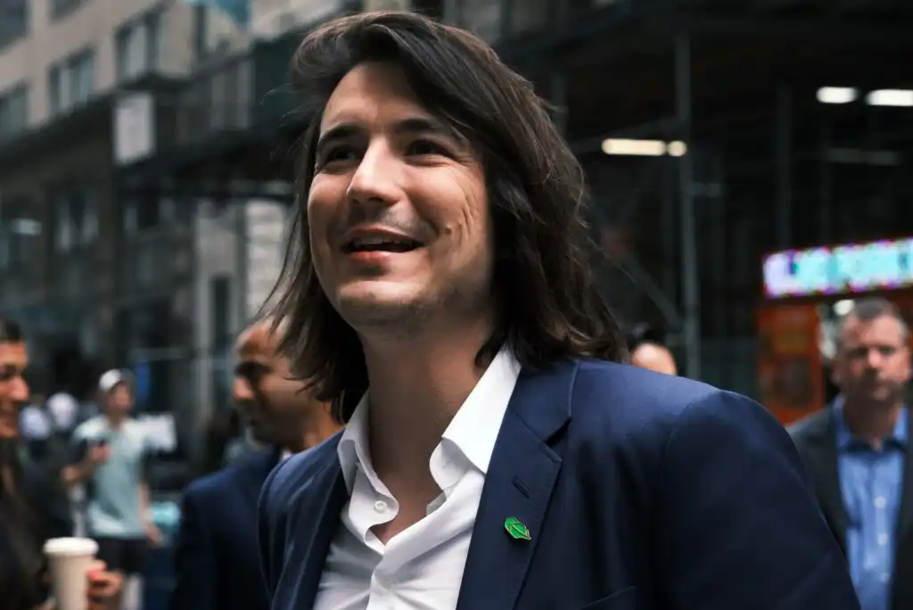 A man with long dark hair, wearing a blue suit and white shirt, smiles while standing outdoors in a city setting with people and buildings in the background.