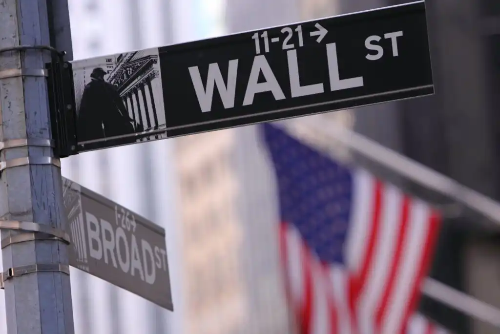 A street sign for Wall Street with a smaller Broad Street sign below it, with a blurred American flag in the background.