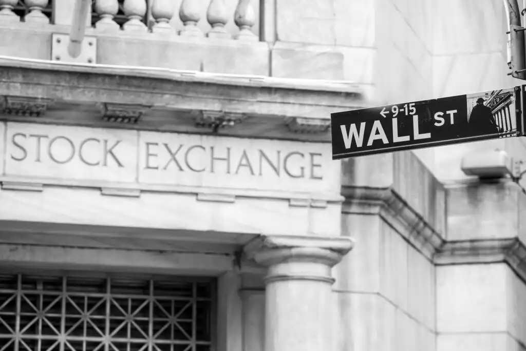 A black and white photo of a Wall Street street sign in front of a building with Stock Exchange engraved on its stone facade.