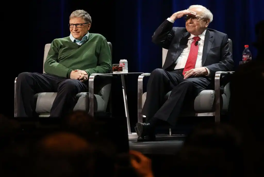Two older men sit on stage in armchairs. One, wearing a green sweater, smiles with hands folded. The other, in a suit and red tie, shades his eyes and looks ahead. Both have drinks on small tables beside them.
