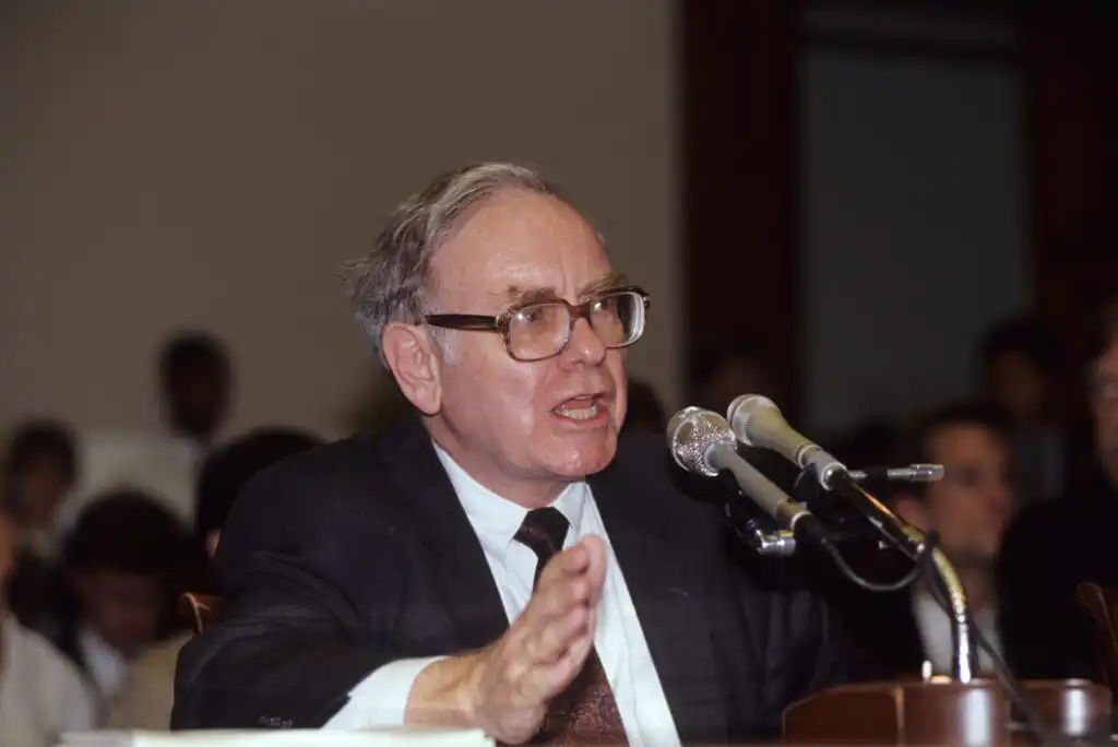 An older man in a suit and glasses speaks into multiple microphones at a formal event, gesturing with his hand, with an audience visible in the background.
