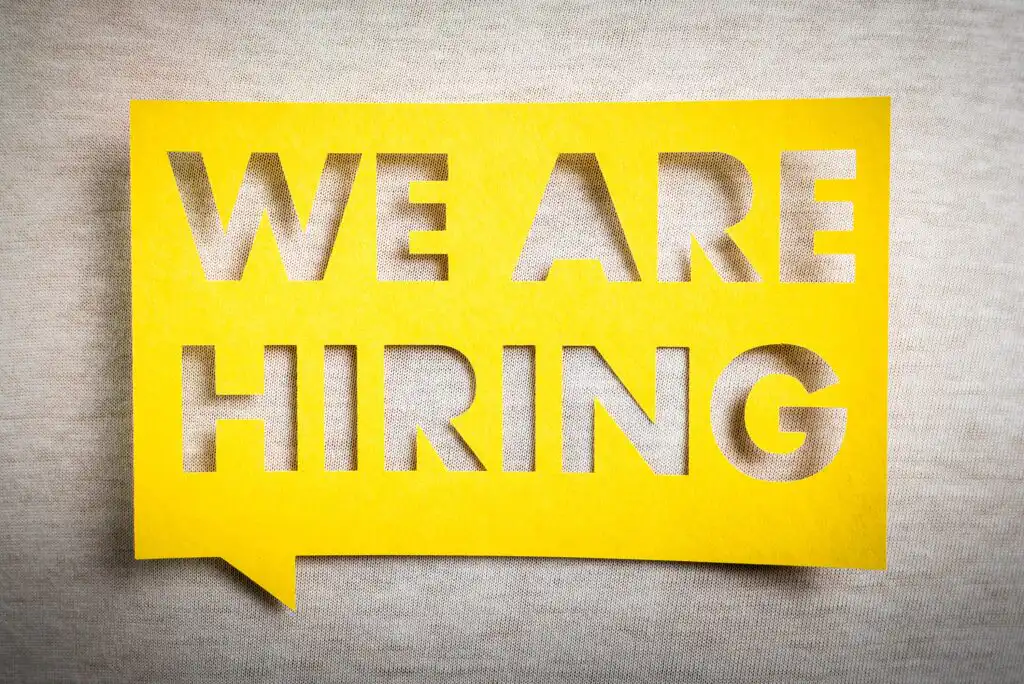 A yellow speech bubble cutout displays the bold text WE ARE HIRING against a light gray fabric background.