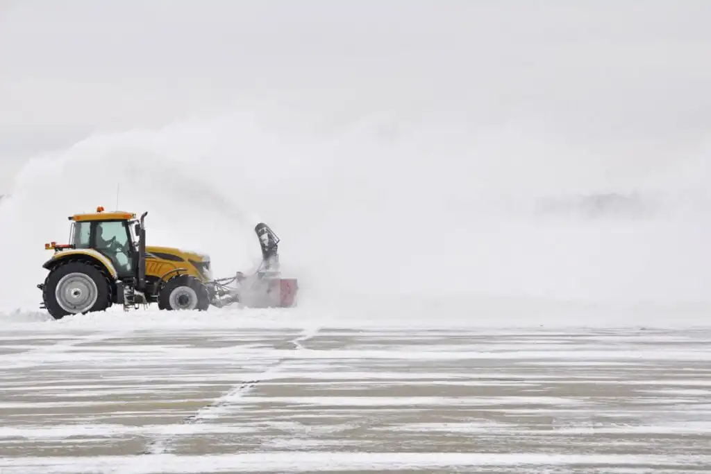 A yellow tractor with a snow blower attachment clears a large amount of snow from an airport runway on a cloudy, overcast day. Snow is blowing high into the air, partially obscuring the background.