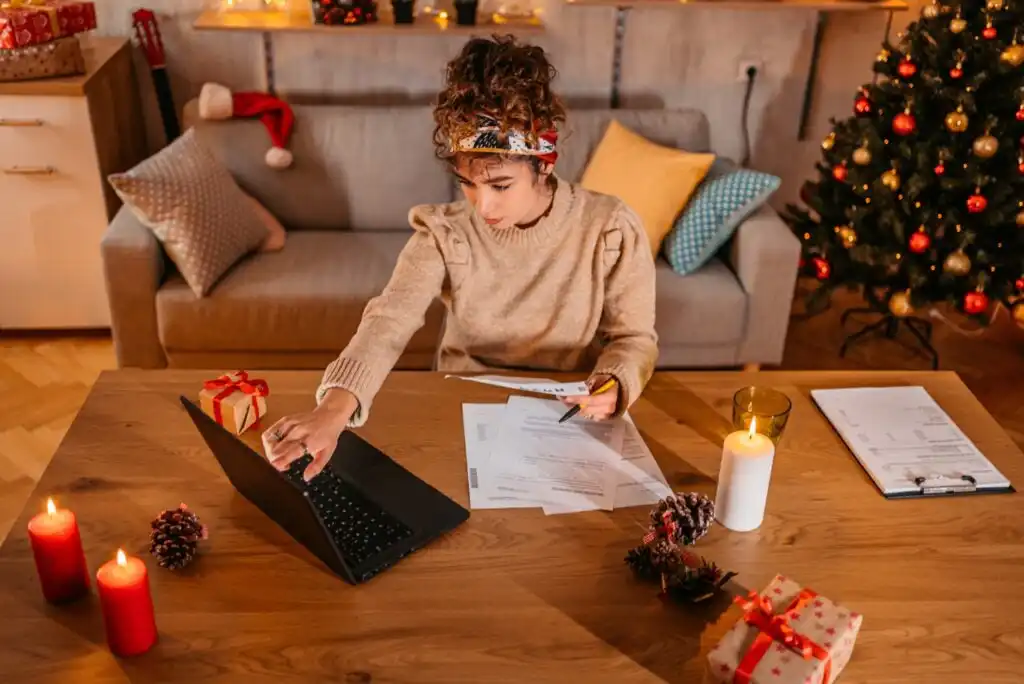 A woman sits at a table with a laptop, papers, and gifts, surrounded by lit candles and holiday decorations, including a Christmas tree and ornaments, working in a cozy living room.